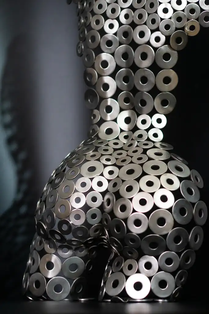 Metal sculpture of a female torso made from hundreds of steel washers, casting a patterned shadow on a dark background.