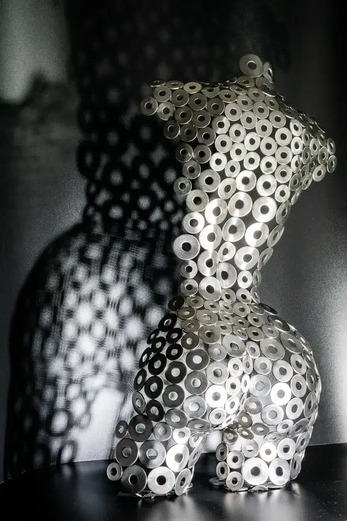 Metal sculpture of a female torso made from hundreds of steel washers, casting a patterned shadow on a dark background.