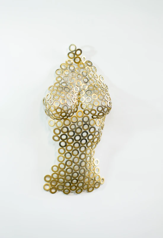 A vertical wall-mounted sculpture depicting a female torso from the neck to the hips. The piece is constructed from numerous small, circular metal washers welded together, creating a mesh-like, transparent effect. The metal has a textured, antiqued gold patina. The sculpture is mounted on a plain white wall, casting soft shadows through its openwork structure. Below it, a corner of a modern gold-framed side table with a dark glass top is visible.